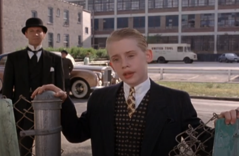 Young boy in formal attire with an adult man in a suit and hat standing behind him near a classic car