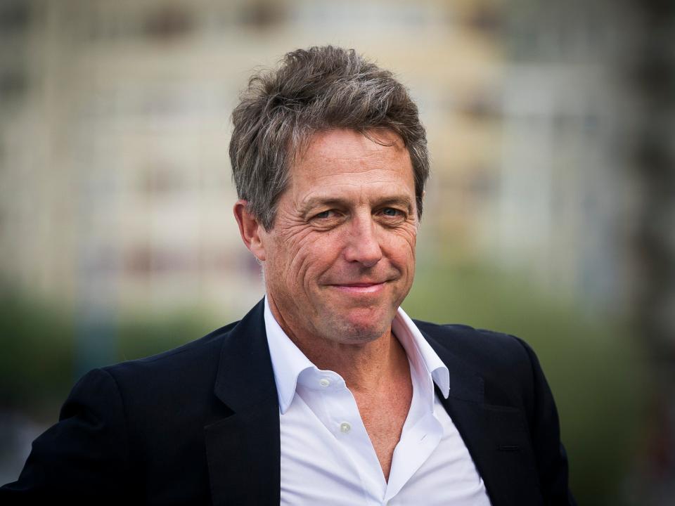 Hugh Grant smiles. He wears a black suit with a white shirt.