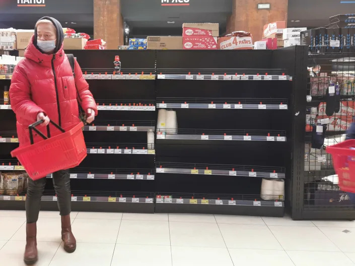 A woman holding a shopping basket stands in front of empty shelves at a supermarket. 