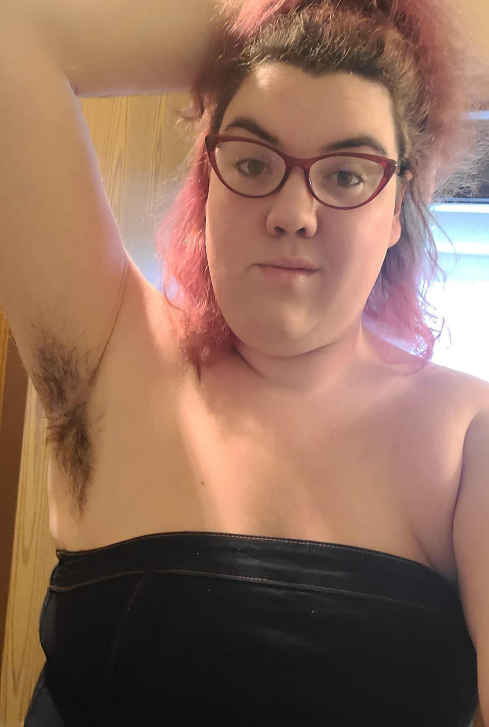 Cassandra Sunshine wearing a black boob tube top and red spectacles while holding one arm behind her head and exposing her underarm hair