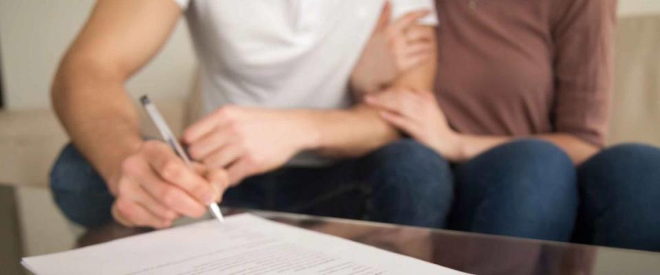 Close up of signing loan agreement, couple sitting on sofa, male hand with a pen setting signature, taking bank credit with easy terms of payment and low interest rate for purchasing property