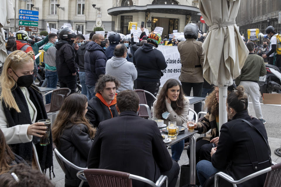 People sit around a table in a sidewalk bar as delivery riders protest behind them outside the Spanish parliament in Madrid, Wednesday March 3, 2021. TFood delivery workers have staged protests across Spain, urging the government to approve a promised law granting them the right to choose between being company staff or self-employed. Media reports said more than 2,000 delivery riders gathered to protest in at least 10 Spanish cities on Wednesday. (AP Photo/Paul White)