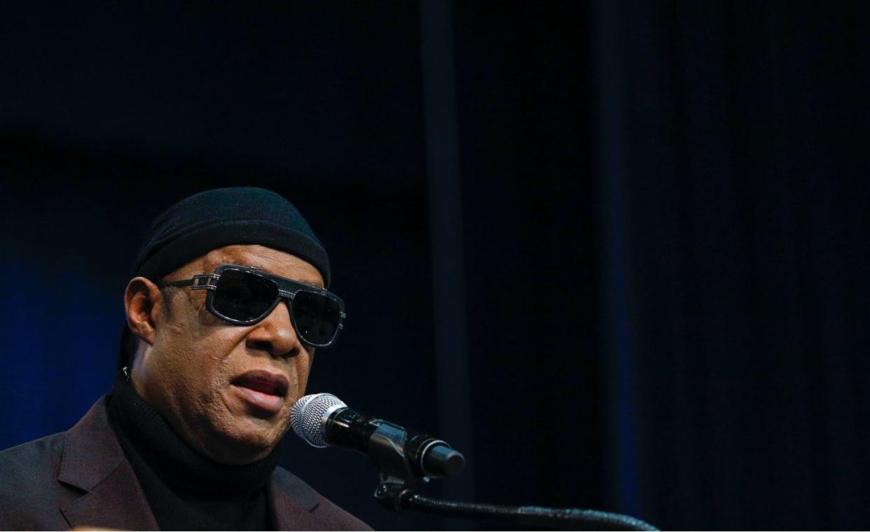 Musician Stevie Wonder speaks into a microphone with his signature sunglasses and a dark maroon suit