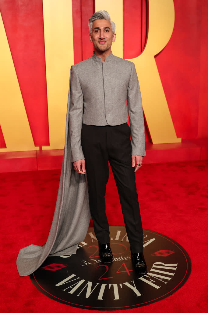 A person in a grey high-neck top with a long flowing train, black trousers, and polished shoes stands on the Vanity Fair red carpet