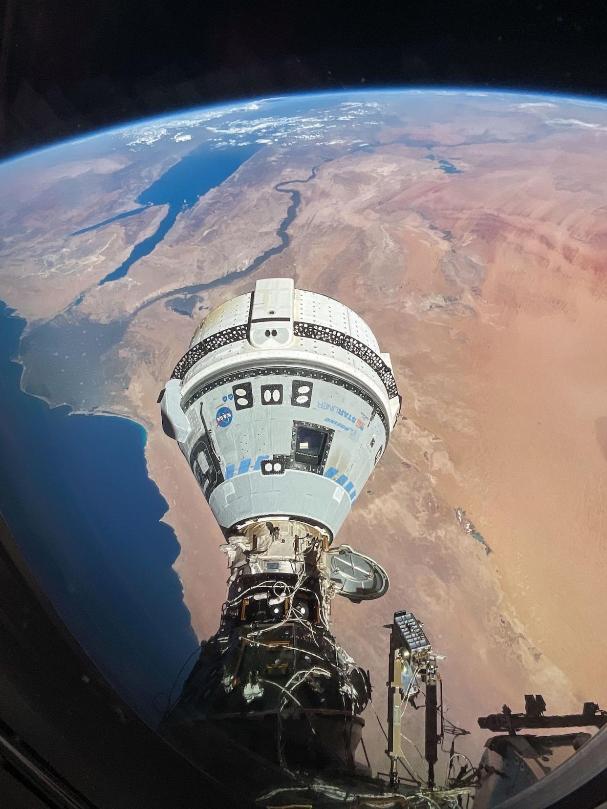 boeing starliner teardrop-shaped spaceship docked to a large wire-covered tube off the side of the space station above earth with a brown and red sandy continent stretching below