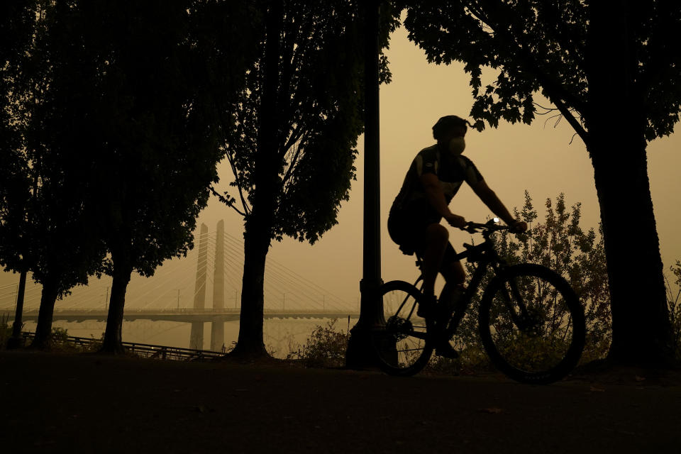 FILE - A person rides a bike along the Willamette River as smoke from wildfires partially obscures the Tilikum Crossing Bridge, Saturday, Sept. 12, 2020, in Portland, Ore. One of North America’s top cycling cities, Portland quadrupled its daily number of bicycle commuters from 2000 to 2019, expanding bike lanes and trails, rerouting car traffic and reducing speed limits — a common mix among the more ambitious pro-bike cities. The cyclist injury rate was halved. (AP Photo/John Locher, File)