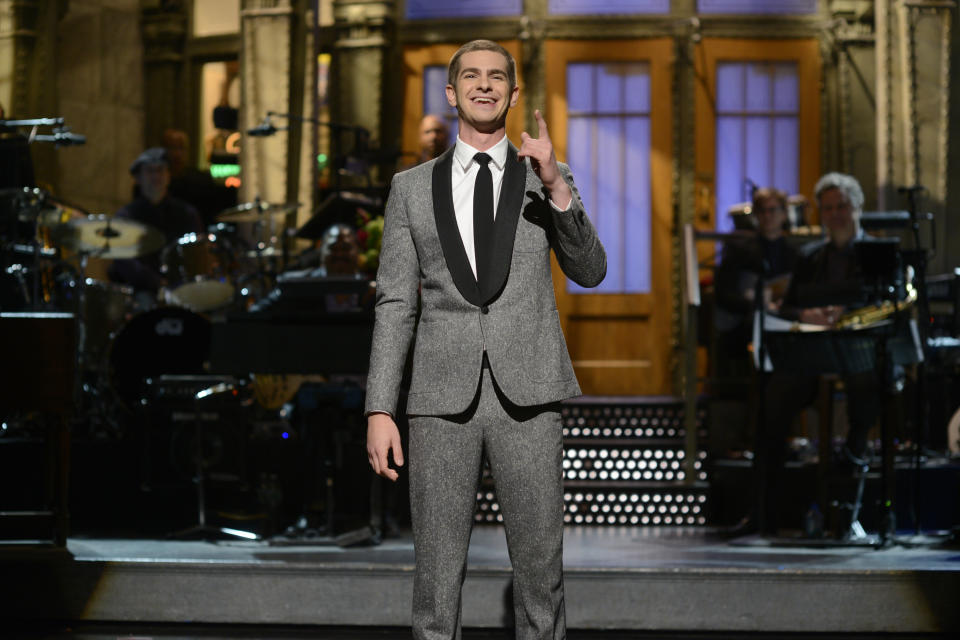 Man in a suit smiling and waving on a stage with a band in the background