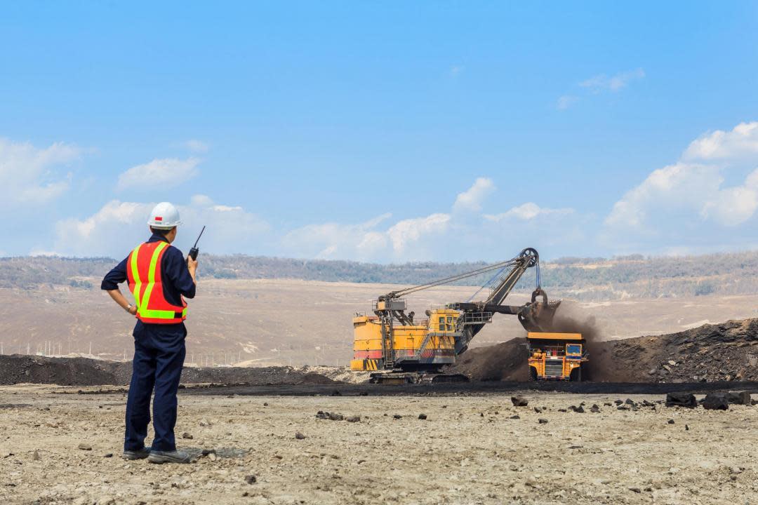 A worker with a high-vis vest and a hard hat holds a walkie talkie while a person operates machinery in the distance.