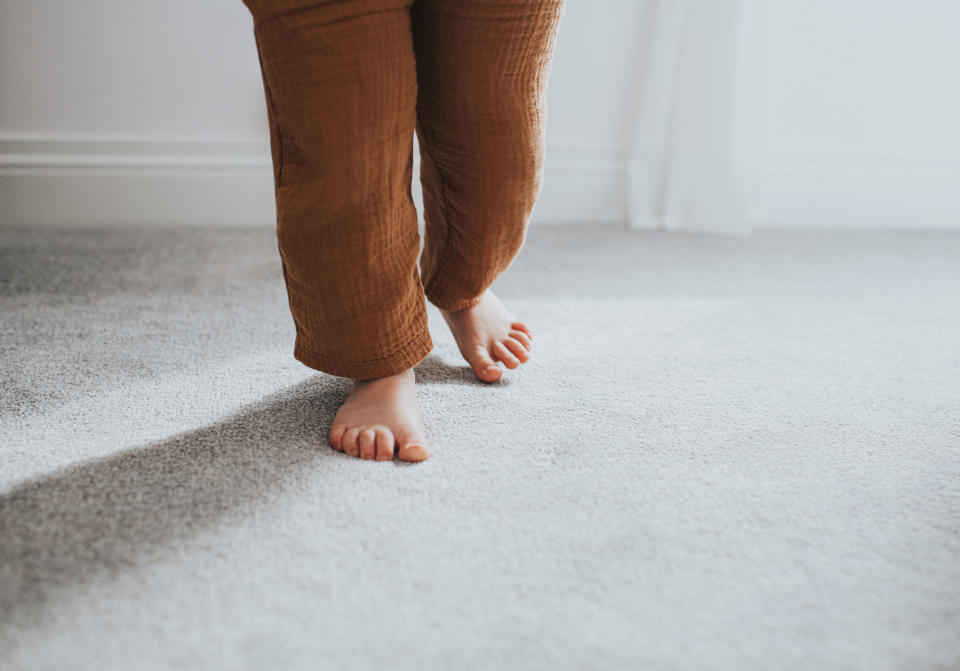 Close-up of a child's feet walking on a soft, gray carpet indoors. The child's legs are partially visible, wearing loose-fitting pants