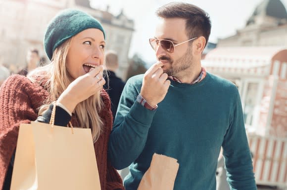 A woman and a man snack on something in a paper bag while walking outdoors.