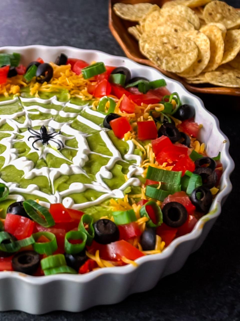 Spooky 8-Layer Dip has refried beans, sour cream mixed with taco seasoning, salsa, guacamole, cheese, tomatoes, black olives, and green onions.