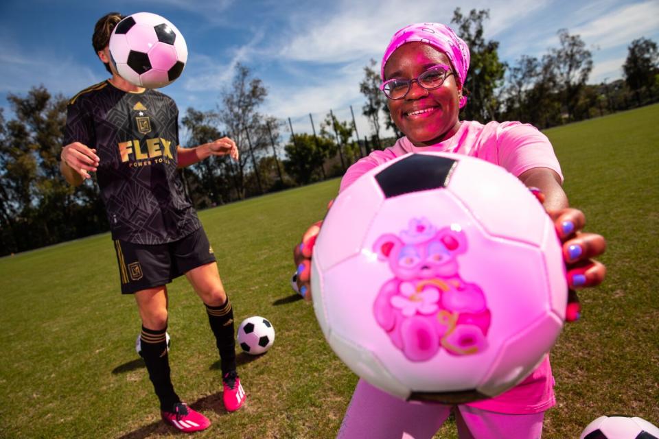 A woman in pink shows off a soccer ball, with a cute image, as a soccer ball obscures a player's face in the background.