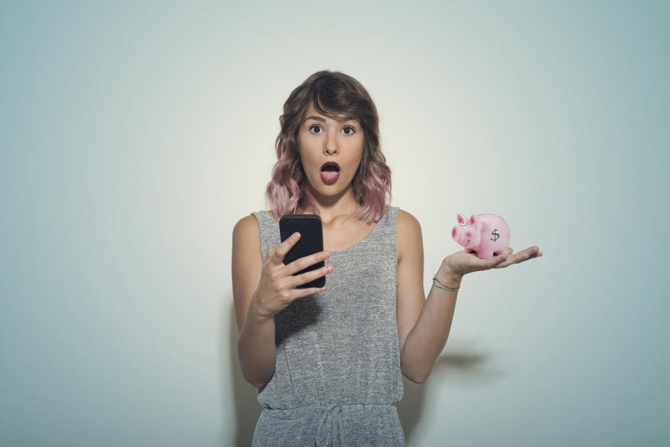 Pictured: A shocked young woman holding a phone and a piggy bank. Image: Getty