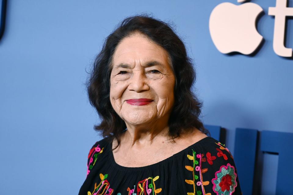 dolores huerta smiles at the camera while standing in front of a blue background, she wears a black shirt embroidered with a multicolor pattern