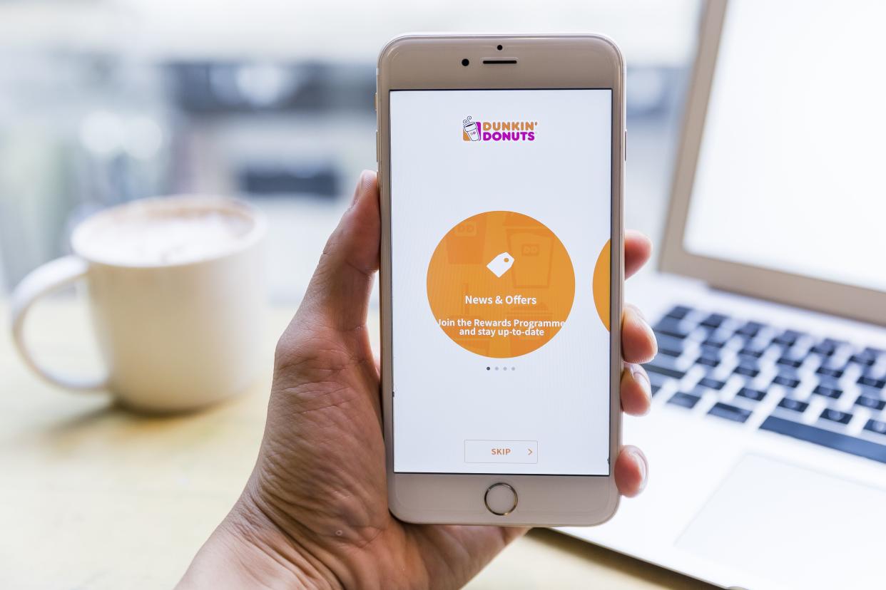 A person holding a smartphone with the Dunkin' app pulled up. In the background is a cup of coffee and a laptop.