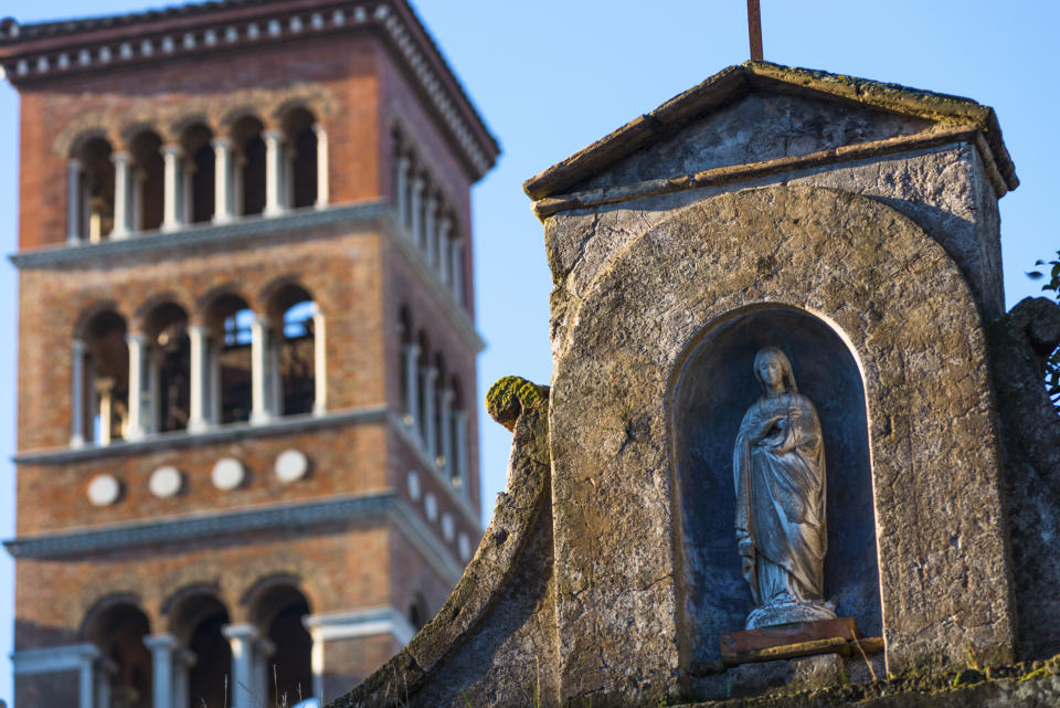 Statue of the Virgin Mary in an alcove of a stone structure with an Italian bell tower in the background