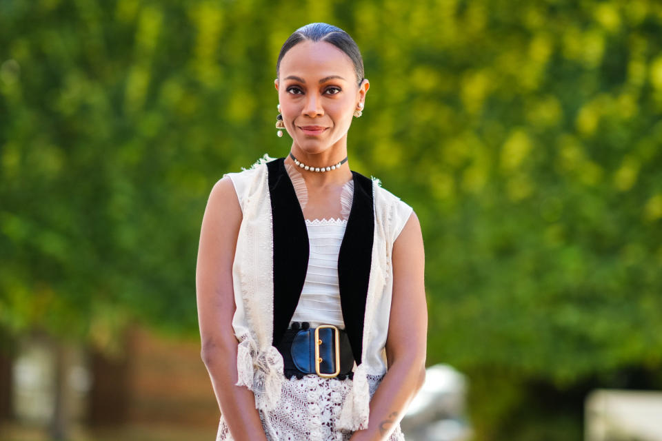 Zoe Saldana wears gold and pearls pendant earrings, a black with white pearls necklace, a white shoulder-off top, a white latte with black borders gilet, a black shiny leather large belt, white lace print pattern midi skirt, gold bracelets, gold rings