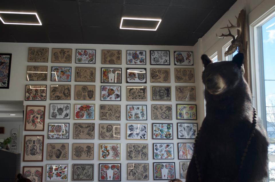 Sebastian the bear stands in front of a wall of tattoo designs at Spilt Milk Mooncusser Tattoo in West Yarmouth.