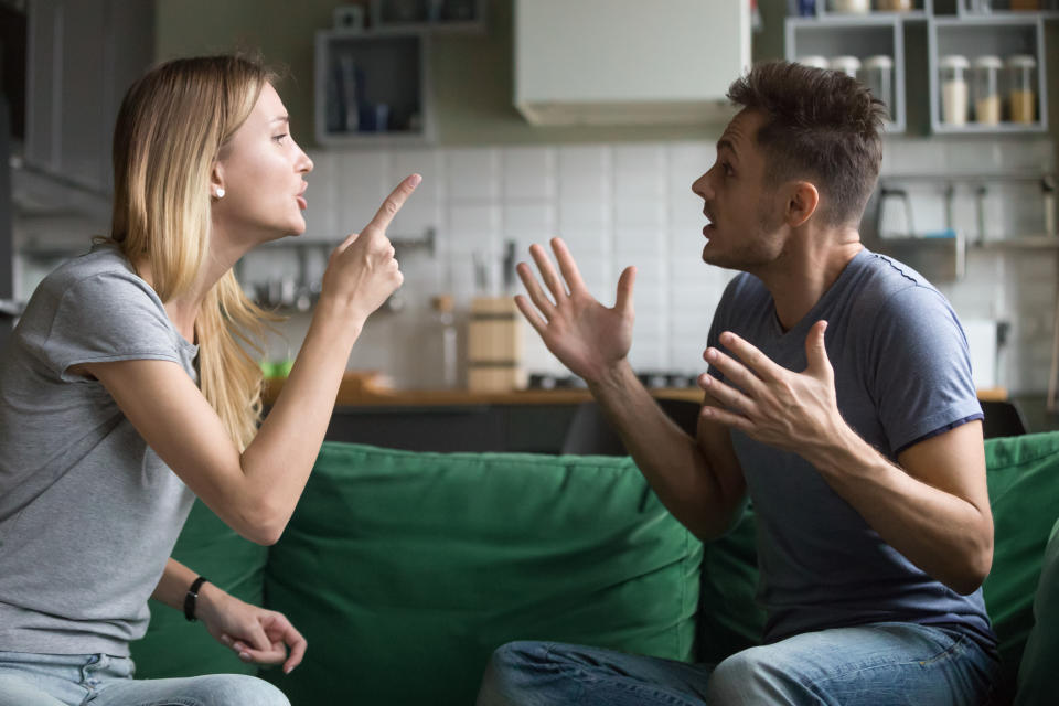 Angry husband and wife arguing on a green couch. Both are wearing grey shirts and blue jeans.