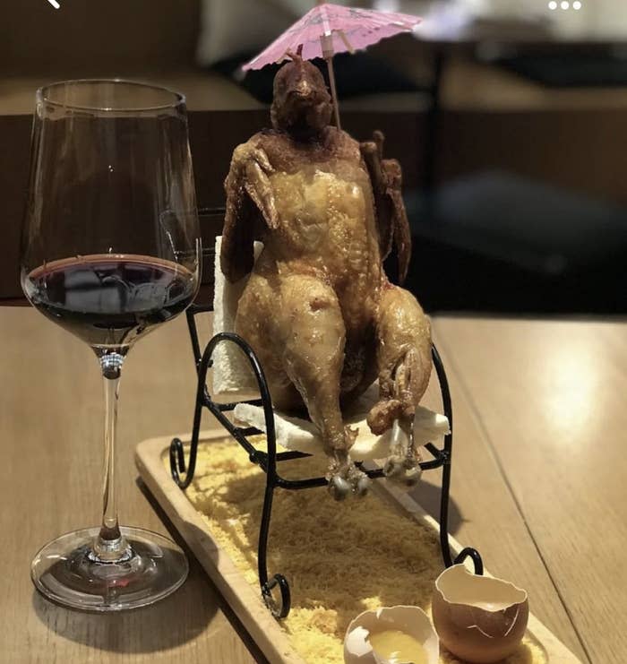 A cooked chicken placed on a small chair and holding an umbrella, making it look like a tiny person