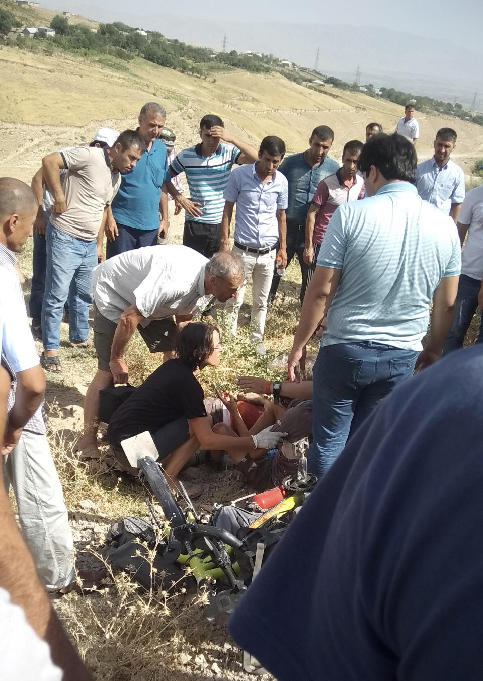 In this photo taken on Sunday, July 29, 2018, people help a wounded cyclist, next to a body of killed cyclist, where four tourists were killed when a car rammed into a group of foreigners on bicycles, south of the capital of Dushanbe, Tajikistan. The Islamic State group on Tuesday claimed responsibility for a car-and-knife attack on Western tourists cycling in Tajikistan that killed two Americans and two Europeans. (AP Photo/Zuly Rahmatova)