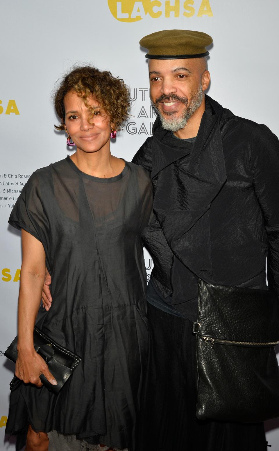 Two people posing together; person on left in a draped dress with clutch, person on right in layered attire with a cap and shoulder bag