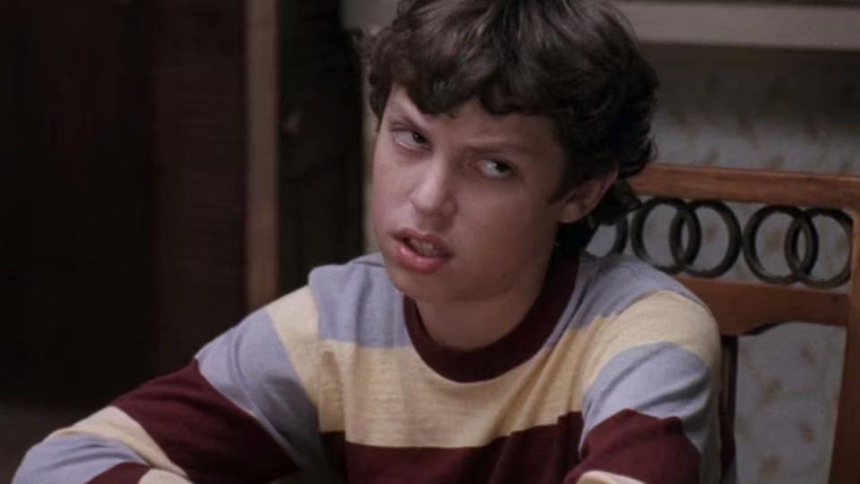 A person sitting at a table with a skeptical expression, wearing a long-sleeve shirt with horizontal stripes