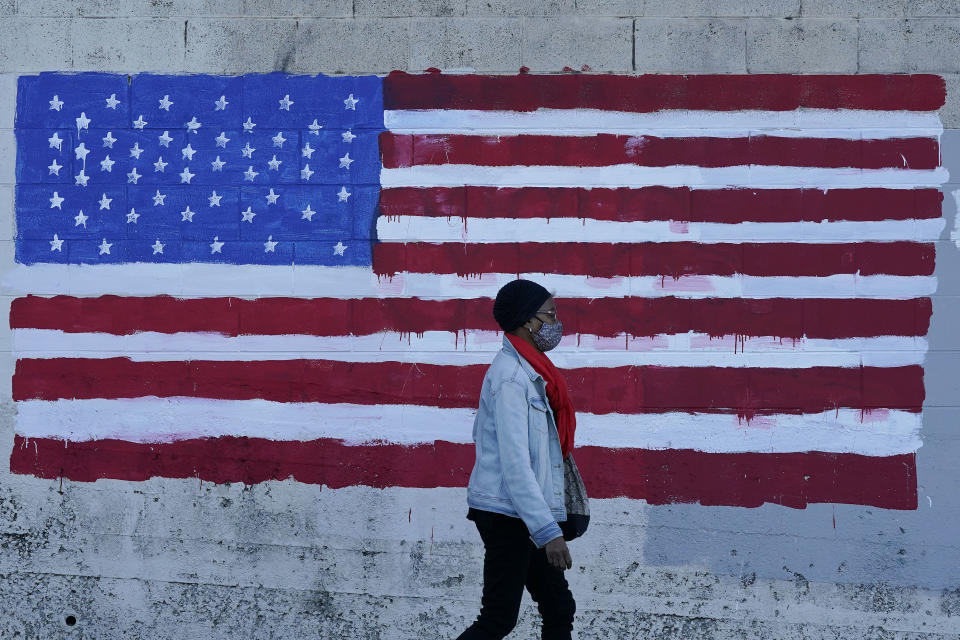 FILE - In this Nov. 16, 2020, file photo, a woman wears a mask while walking past an American flag painted on a wall during the coronavirus outbreak in San Francisco. A deadly rise in COVID-19 infections is forcing state and local officials to adjust their blueprints for fighting a virus that is threatening to overwhelm health care systems. (AP Photo/Jeff Chiu, File)
