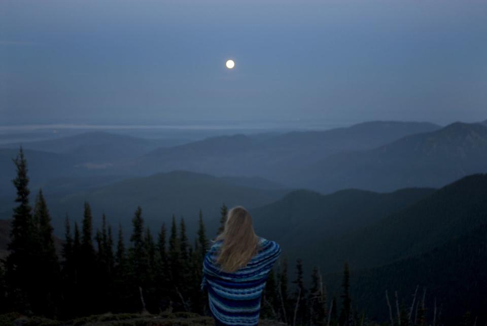 woman wrapped in blanket looking at mountainous landscape with full moon, rear view