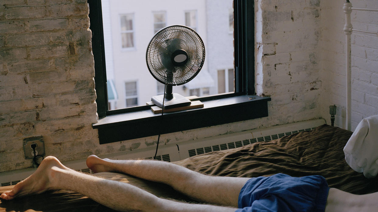  man lying on bed face-down with window open and fan on 