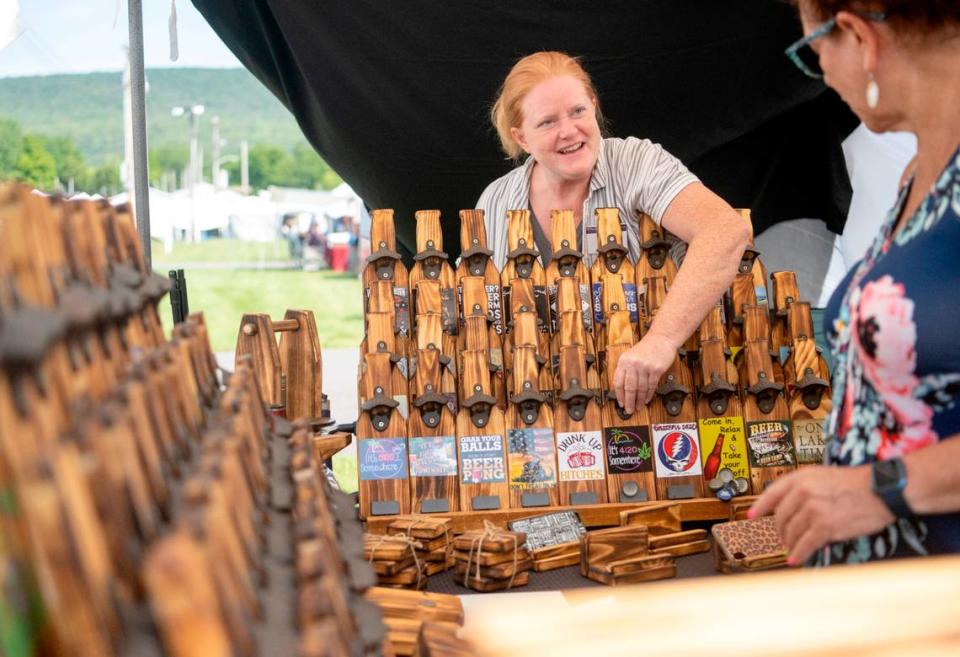 Bonni Oliver talks about her wooden bottle cap openers and games that she makes out of pine as a customer browses her booth at the People’s Choice Festival on Friday, July 14, 2023.