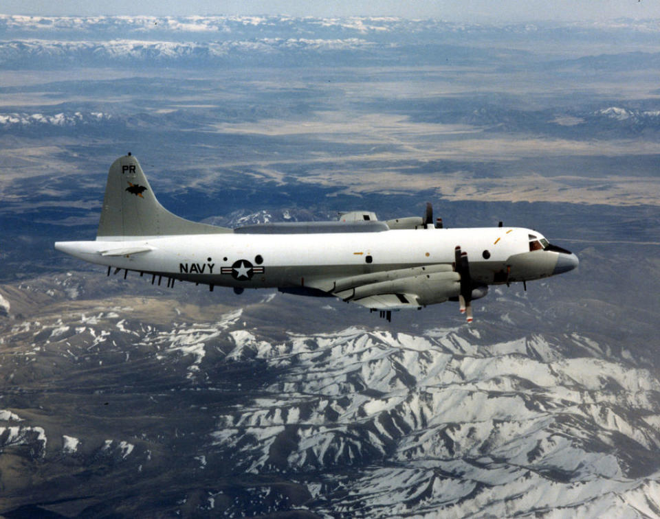 A Navy P-3 Orion aircraft is flying over a mountainous landscape with snow-covered peaks