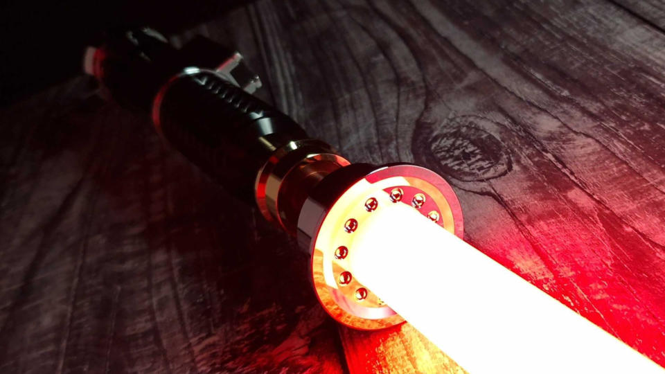 A SabersPro Obi Wan EP3 blade shines red on a wooden surface