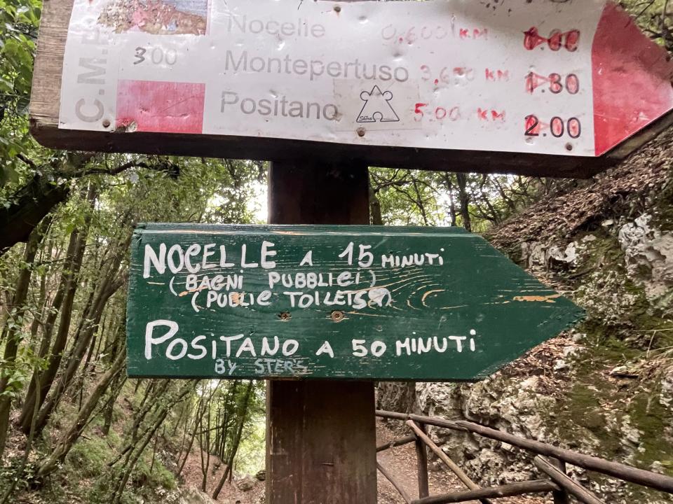 Two signs pointing to the right. The bottom sign reads, in both Italian and English, "Nocelle, 15 minutes (public toilets)" and "Positano 50 minutes by steps."