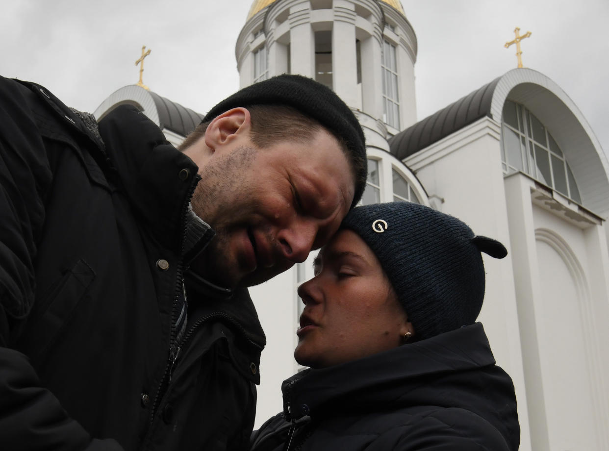 A couple comfort each other next to a man's grave in the shadow of the Church of St. Andrew and All Saints in Bucha.