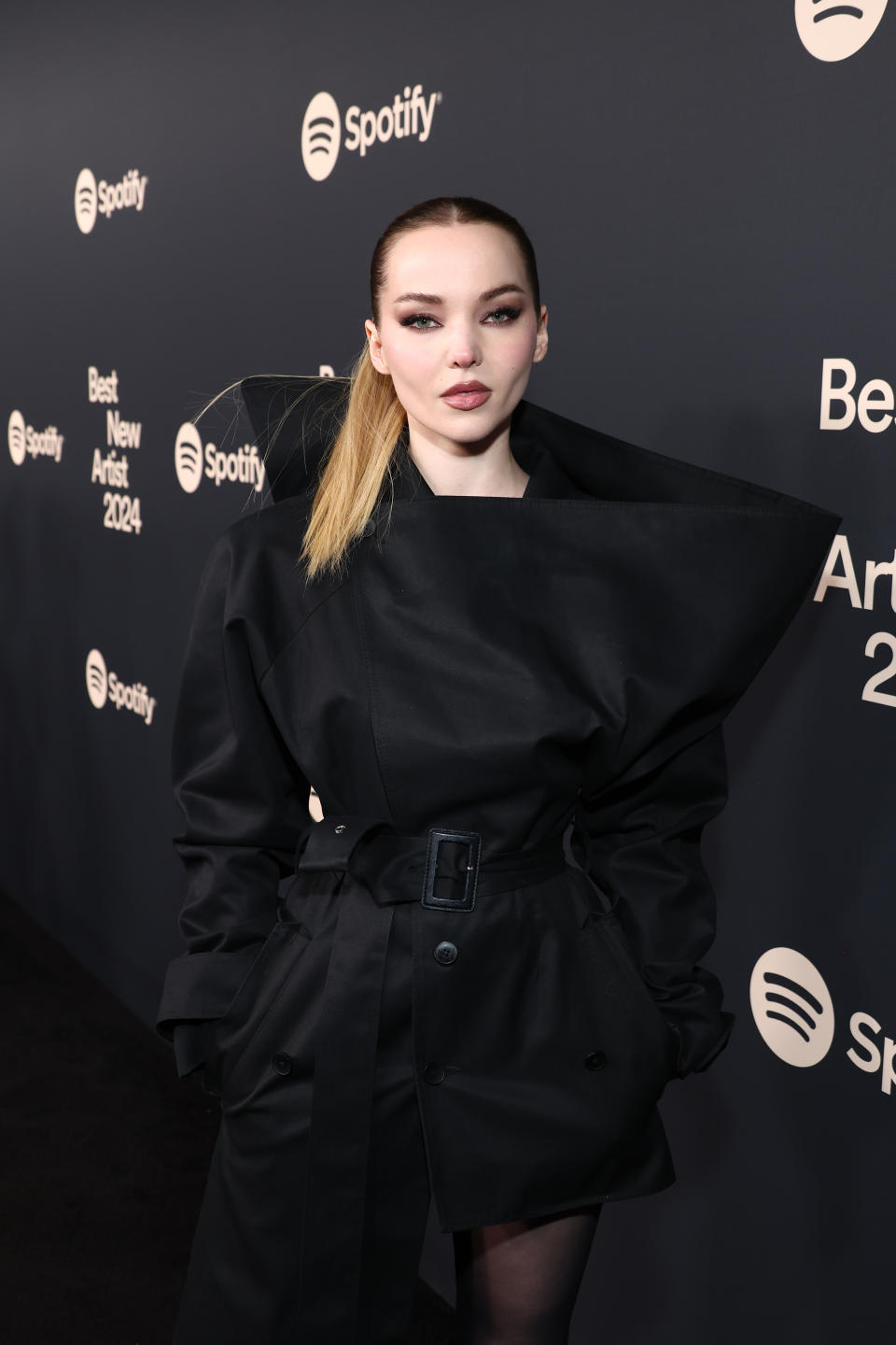 Dove Cameron stands in front of a step and repeat with a multicolor ponytail