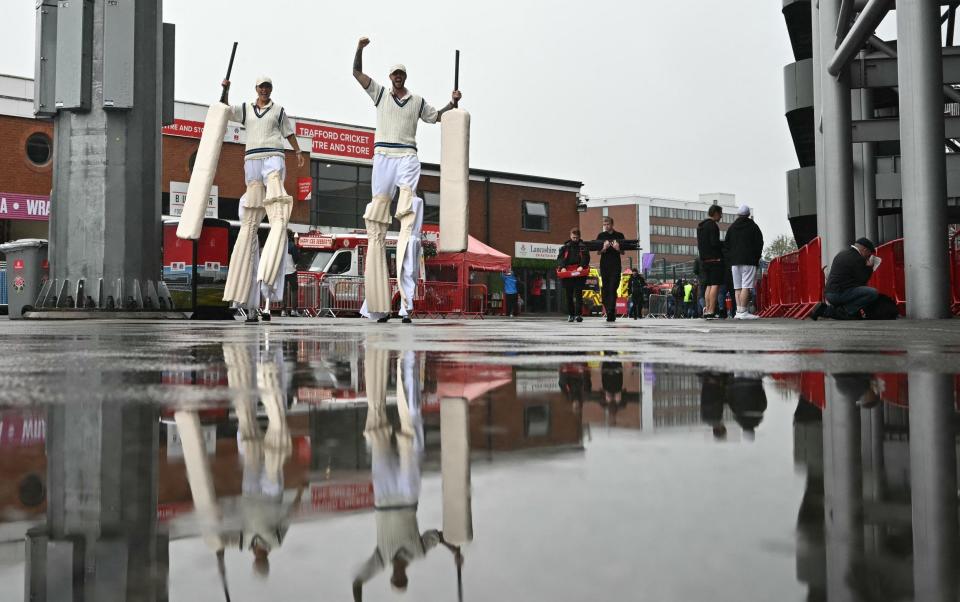 Performers on stilts wearing oversized cricket gear walk through the puddles behind the grandstand as rain falls prior to the start of play on day two of the first Test match between England and Sri Lanka at Old Trafford cricket ground in Manchester, north-west England on August 22, 2024