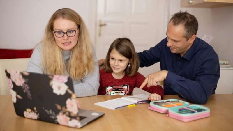 A mother and father sit with their young daughter at a table, looking at an open laptop, with an open book and an open case of colored pencils on the table.