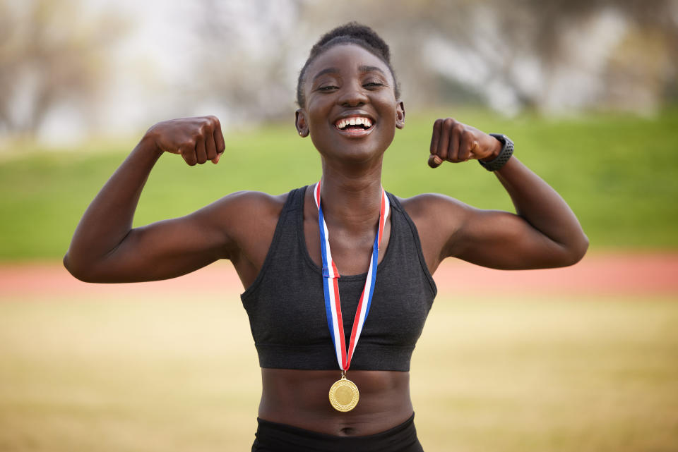 A smiling person proudly flexes their arms while wearing a medal around their neck, standing in an outdoor track field