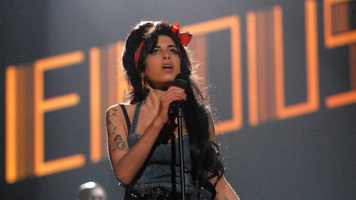 amy winehouse holding a standing microphone and singing as her name is illuminated in the background