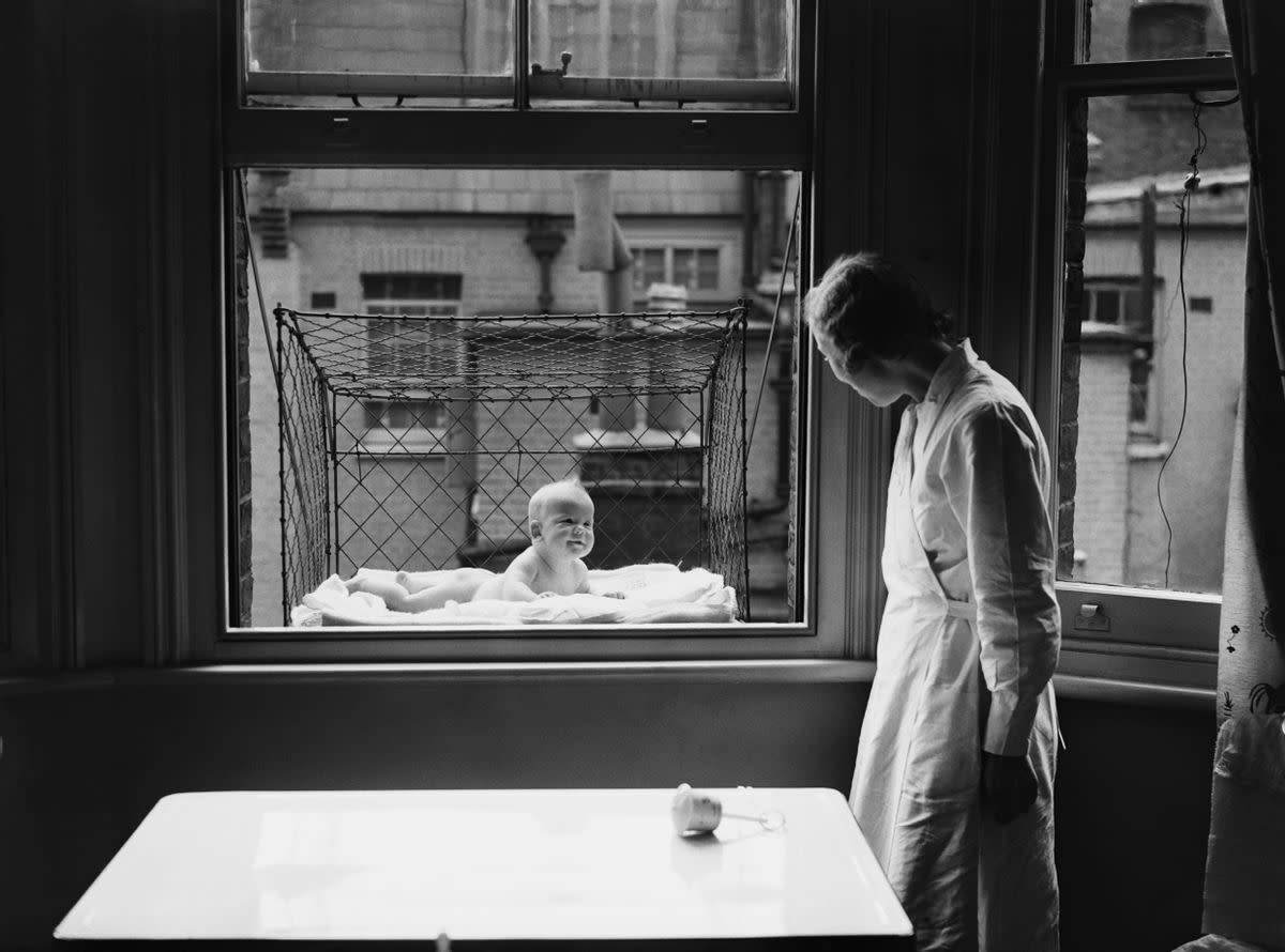 A black and white photo shows a nurse wearing a white coat looking at a baby in a wire cage attached to a window with blankets covering the bottom looking at them. . 