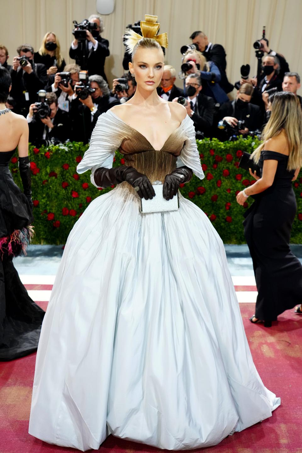 Caroline in an off the shoulder full-skirt white dress with a brown ombre on the bodice, chest, and neckline. The off the shoulder sleeves each her elbows and reveal brown leather opera gloves. She's holding a white clutch and her hair is up in a sculptural bun.