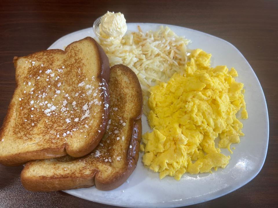 Two slices of French toast with a light dusting of powdered sugar, scrambled eggs, and hash browns on a white platter