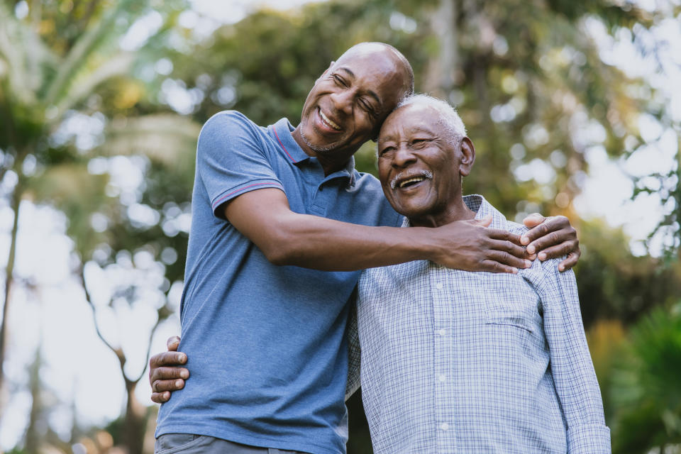 Two elderly men joyfully embrace and smile while standing together outdoors, surrounded by trees and greenery