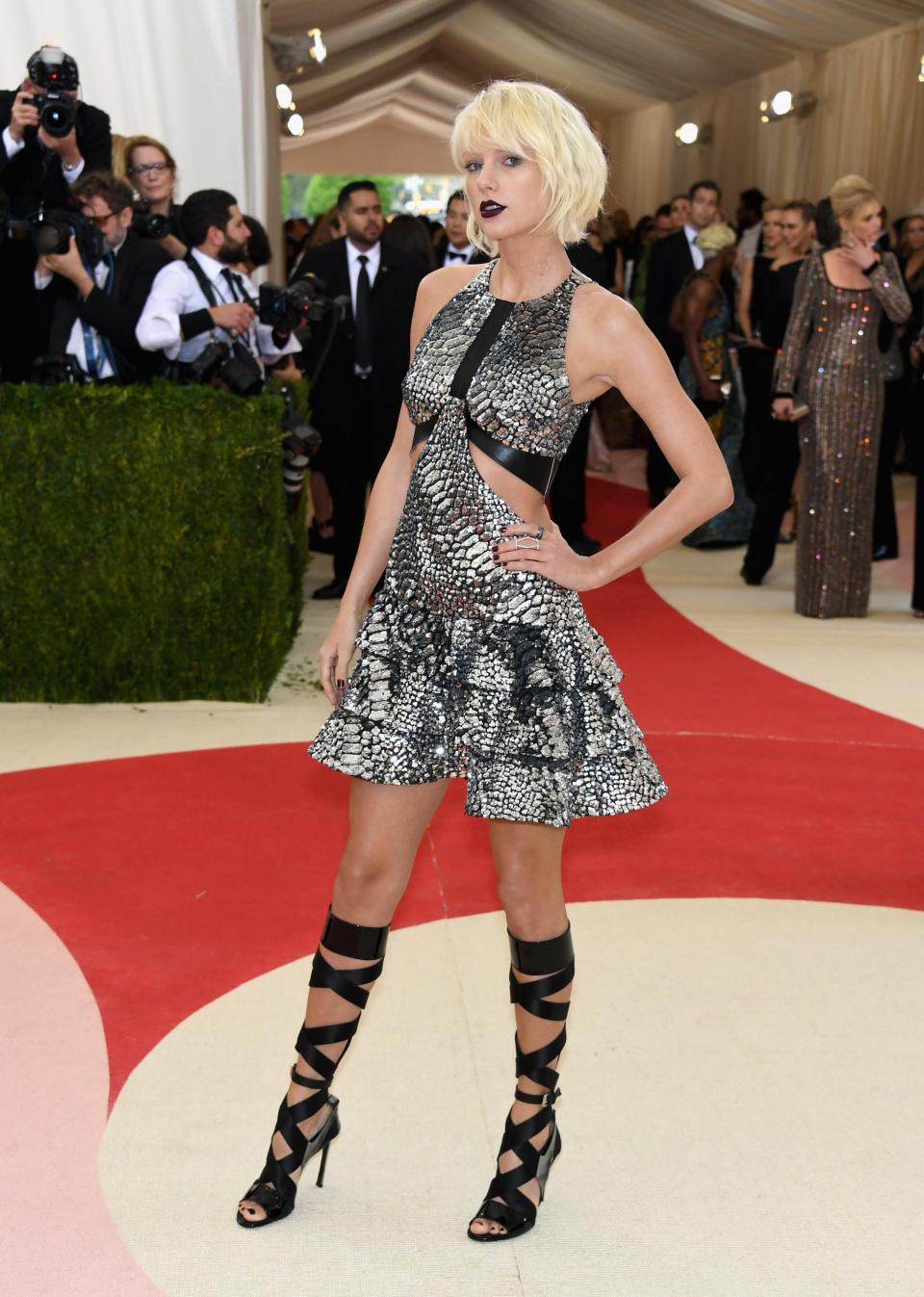 Taylor Swift poses at the Met Gala with bleached hair and wearing a short, flared metallic dress, dark lipstick, and strappy black heels.