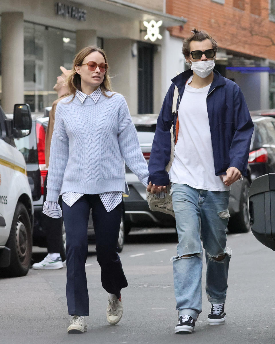 A paparazzi photo of Olivia Wilde and Harry Styles walking on the street hand in hand wearing casual clothes, jeans and sneakers, he's wearing a mask