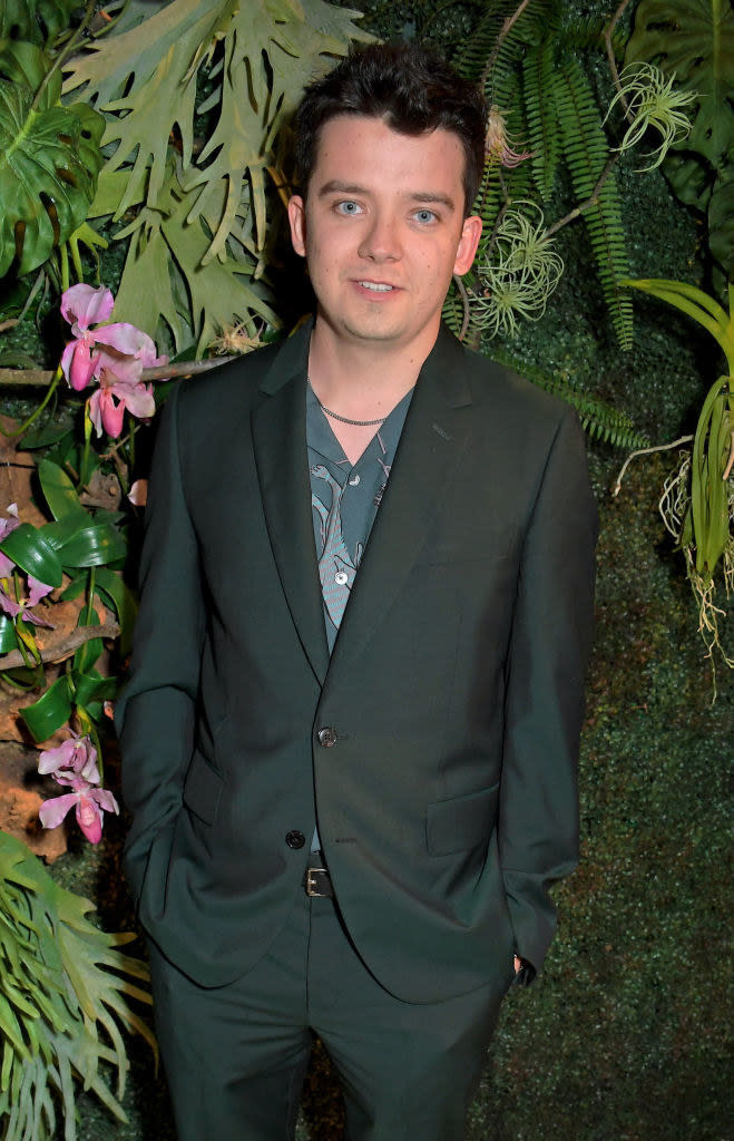 Asa Butterfield standing by greenery and wearing a suit