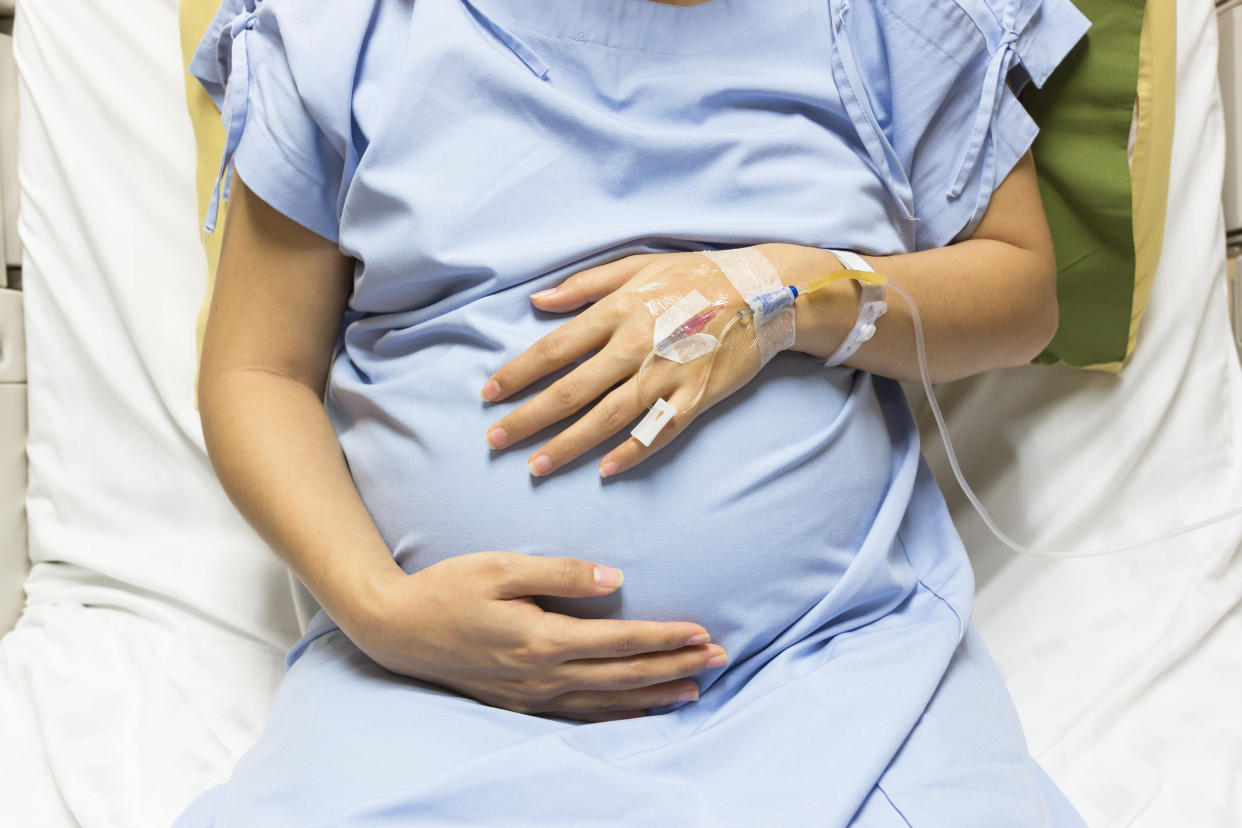 A pregnant woman, left hand connected to an IV, in a hospital bed cradles her belly with her right hand/