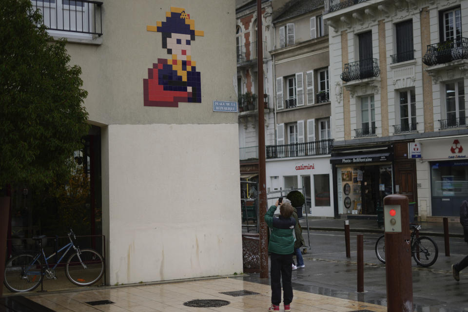 A boy takes a photo of a mosaic by French artist Invader depicting Napoleon, on a street of Fontainebleau, south of Paris, Thursday, Feb. 29, 2024. For the Paris Olympics, it could almost have been a new sport: Score points by scouring France's capital for mosaics that a mystery artist who calls himself "Invader" has cemented to walls across the city, across the world and even had carried aloft to the International Space Station. (AP Photo/Thibault Camus)