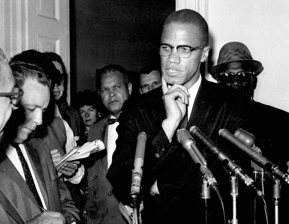 A tall man wearing glasses and speaking to a crowd of reporters